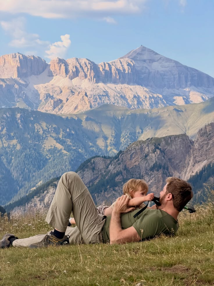 A dad lying back in alpine grass with his toddler on his chest, mountains behind them.