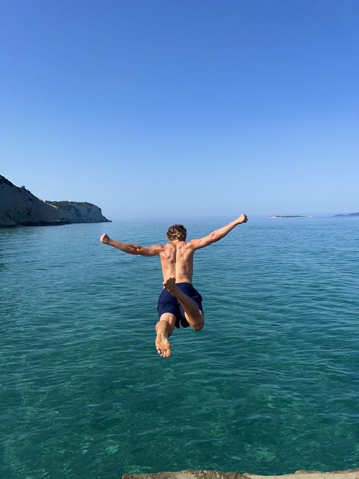 A man leaping off a rock ledge into clear blue sea, arms spread wide.