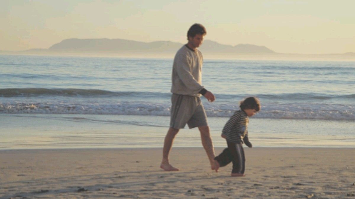 A dad walking barefoot along the beach at sunset beside his toddler.