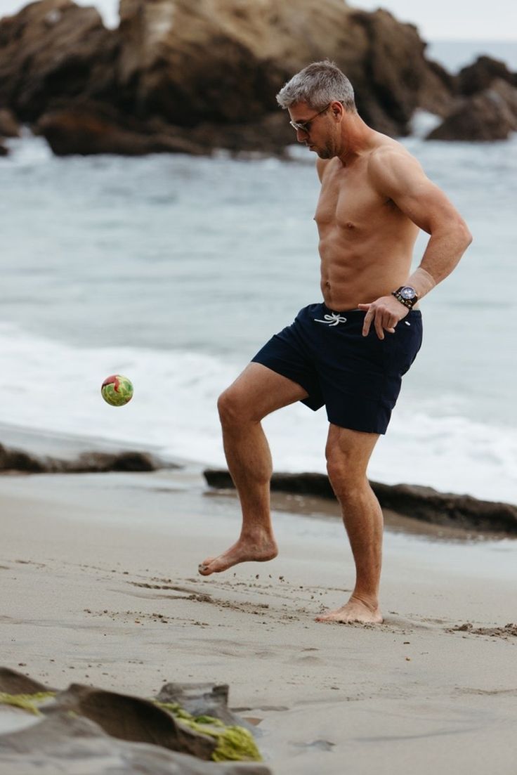 A fit, grey haired man in his forties juggling a ball barefoot on the beach.